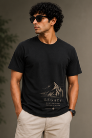 A man wearing a black short-sleeve graphic t-shirt with a mountain design and the text 'LEGACY BUILT ON FAITH GROWN IN ACTION', paired with beige pants, set against a neutral gray background.