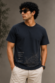 A man wearing a dark navy blue crewneck t-shirt with a subtle compass graphic and 'ORIGINS' text on the front, paired with beige trousers, set against a neutral gray background.