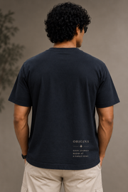 A man viewed from behind wearing a dark navy blue t-shirt with 'ORIGINS' printed in white text on the lower back, paired with beige cargo pants against a neutral gray background.