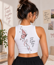 A woman shown from the back wearing a white cropped tank top with a pink butterfly graphic and the phrase 'same girl brighter days' on the back, paired with black shorts in a bright indoor room.